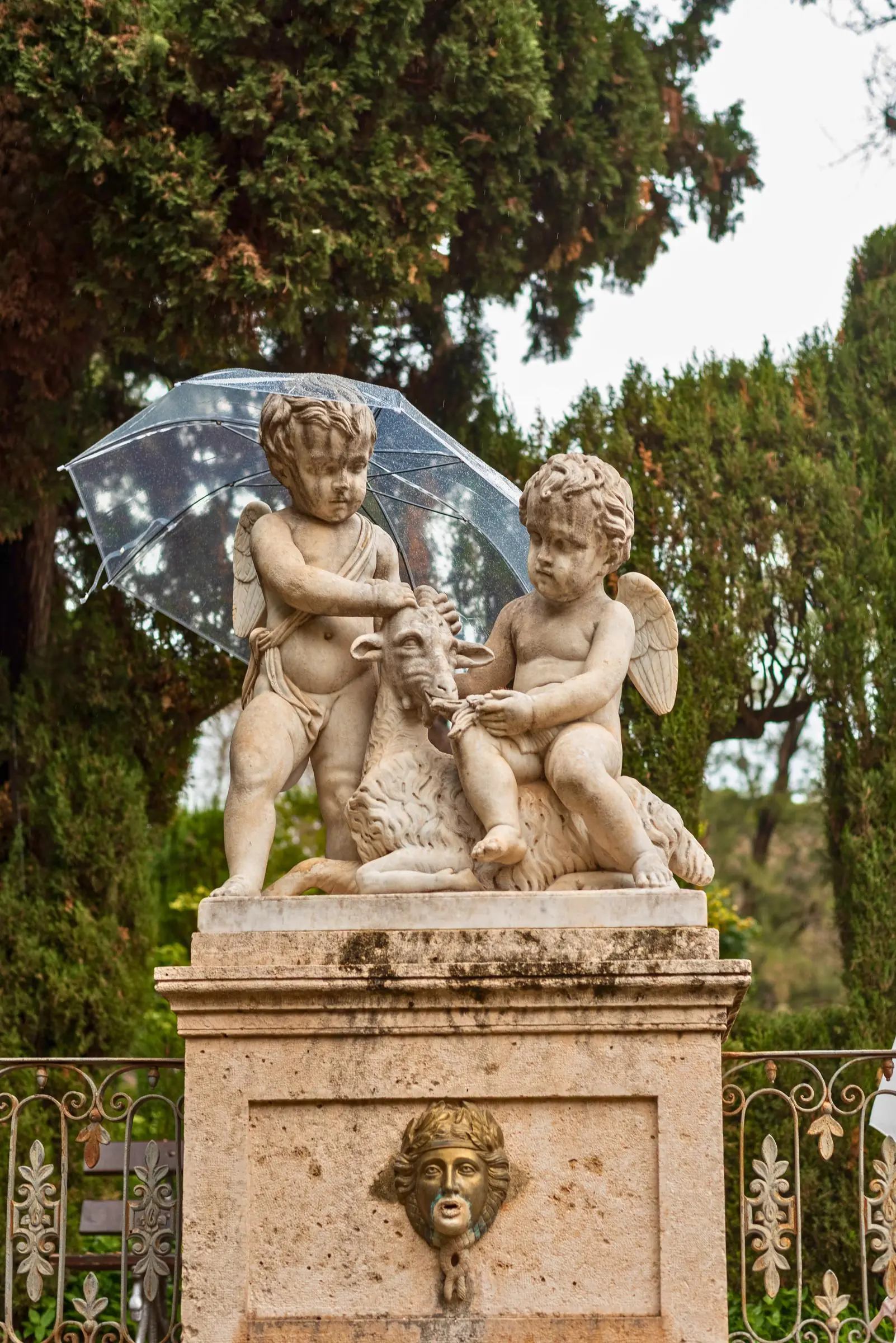 Classical marble statue of two cherubs and a goat shielded from the rain by a clear umbrella